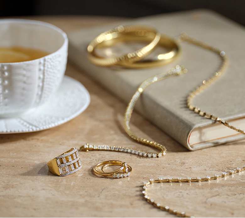 An assortment of lab grown diamond jewelry in yellow gold sitting on a table with a coffee mug and book sitting in frame. Shop lab grown diamonds
