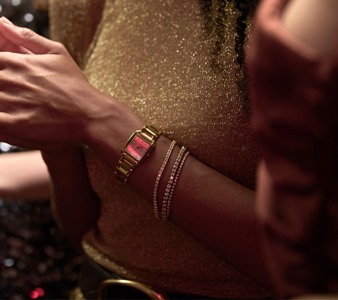 Close-up of an arm wearing a gold-tone rectangular watch and two diamond bracelets. The person is dressed in a glittery gold top, with other people partially visible in the background.