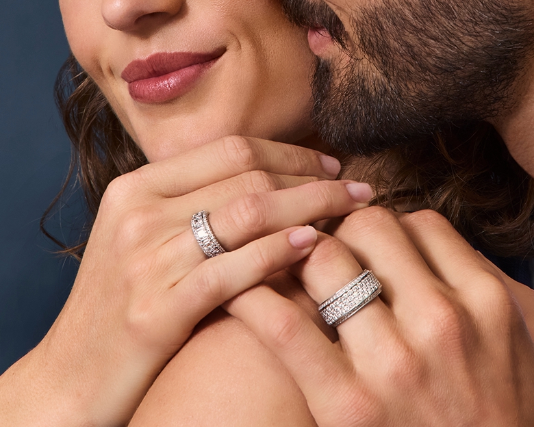 An image of a male and female model from nose down featuring their hands on top of one another's showing their wedding bands.